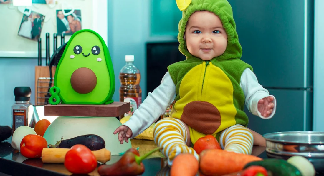 A baby wearing a green hooded outfit sits on a kitchen counter beside fresh vegetables and an avocado-themed lunch box.