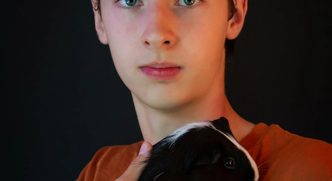 Close-up of a young person in an orange shirt cradling a black and white guinea pig against a dark background