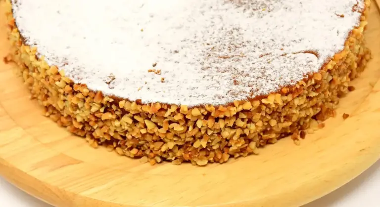Close-up of a round cake with powdered sugar on top and a crust of chopped nuts around the sides, set on a wooden board.
