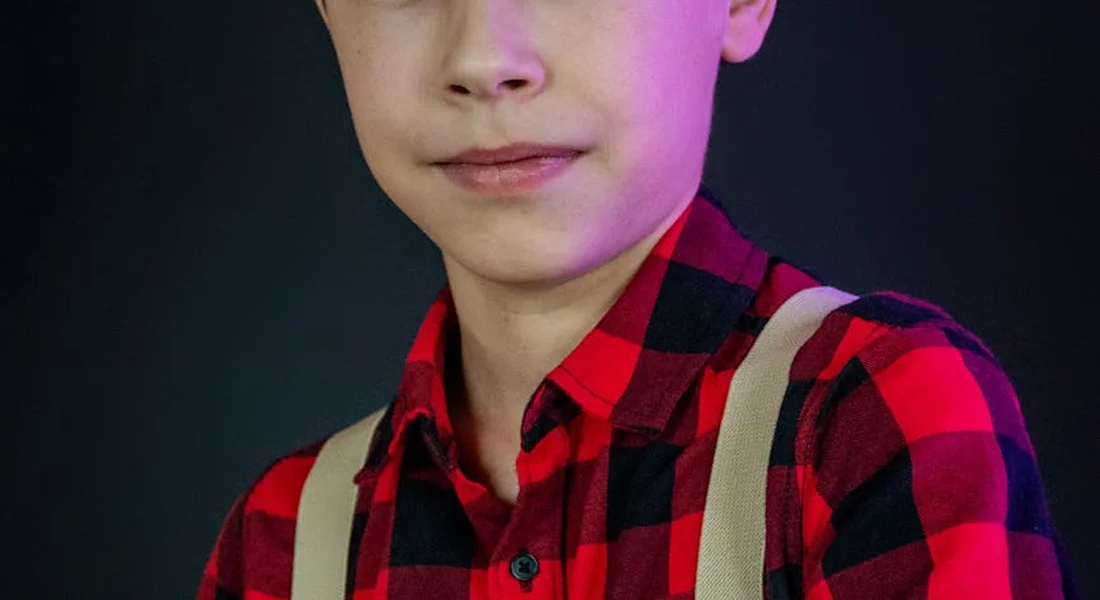 Close-up portrait of a young person wearing a red plaid shirt and beige suspenders against a dark background.
