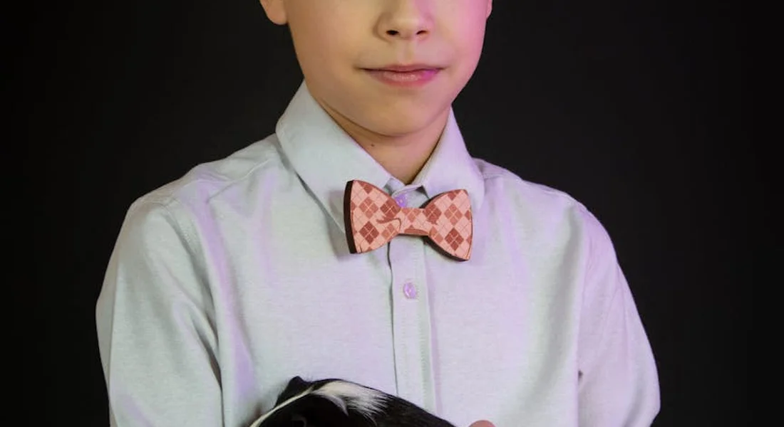 Young person in a light shirt and bow tie holding a guinea pig against a dark background.