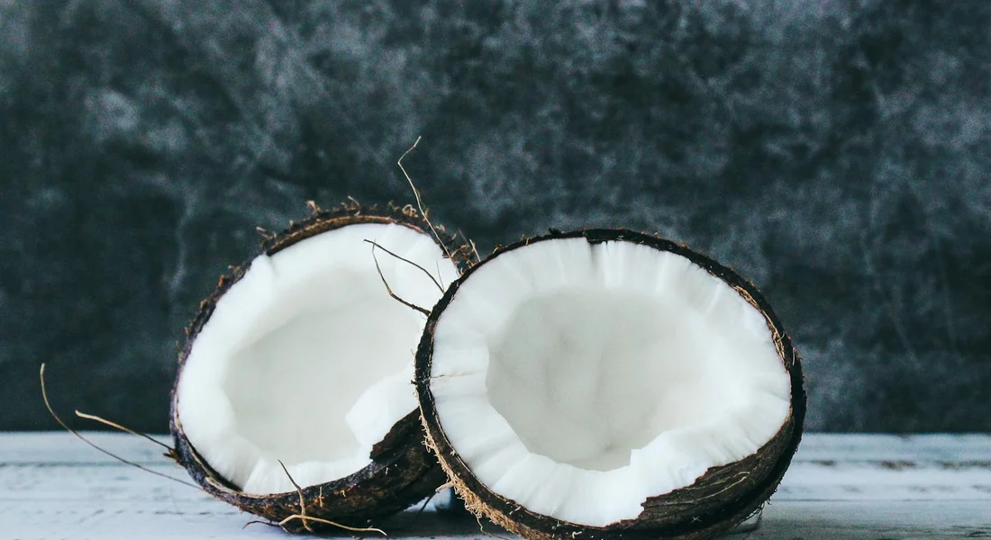 Two halved coconuts with white flesh exposed on a light wooden surface against a dark, textured background