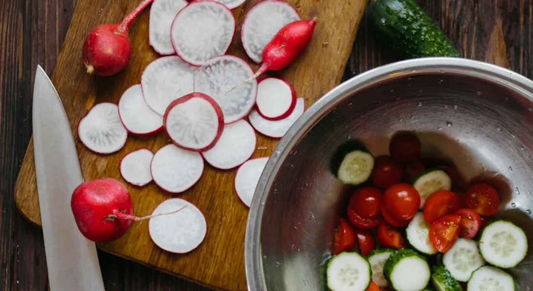 Daikon radish slices on a wooden cutting board, with a knife nearby, and a metal bowl containing cucumber slices and cherry tomato halves.