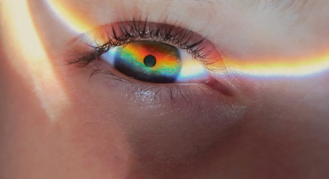 Close-up of an eye with a vivid rainbow light reflection across the eyelid.