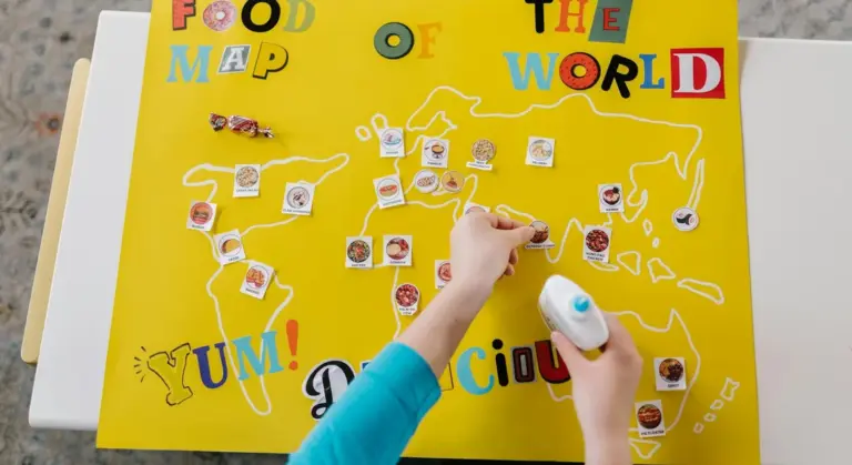 A bright yellow world map collage titled 'FOOD MAP OF THE WORLD' with small photos attached around the map and a hand applying glue from a bottle.