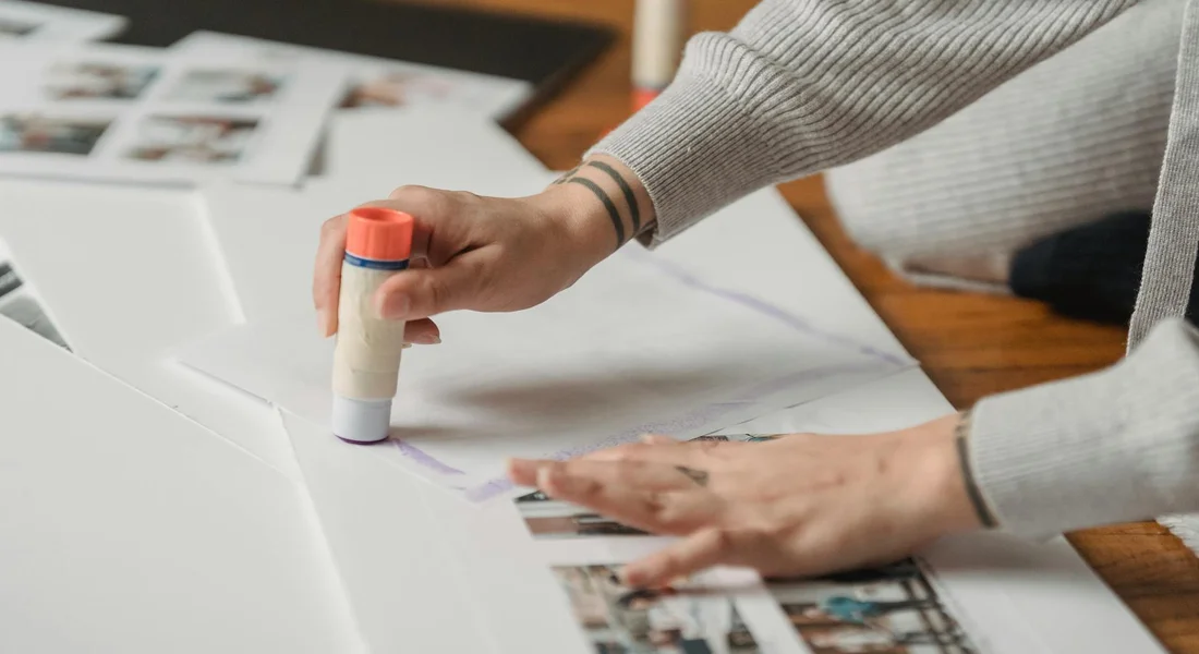 A person uses a glue stick to apply adhesive to photos on a craft project at a desk.
