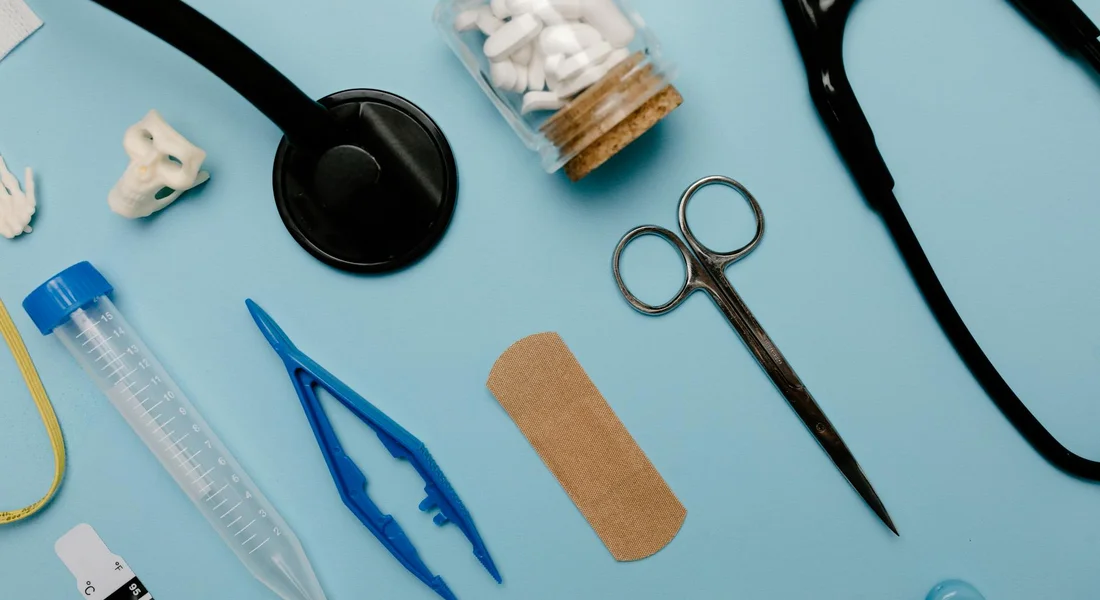 Assorted veterinary tools and medical supplies arranged on a blue background, including a stethoscope, scissors, tweezers, a bandage, a test tube, a pill bottle, and a syringe-like instrument, illustrating supplementary items for advanced guinea pig care.
