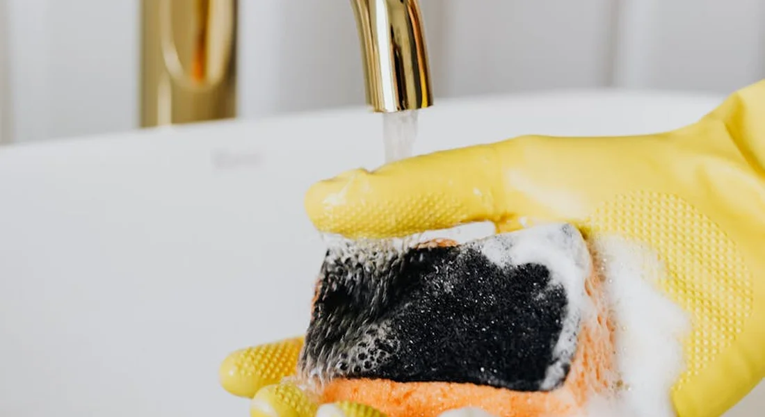 Close-up of a yellow rubber glove holding a soapy sponge under a running faucet