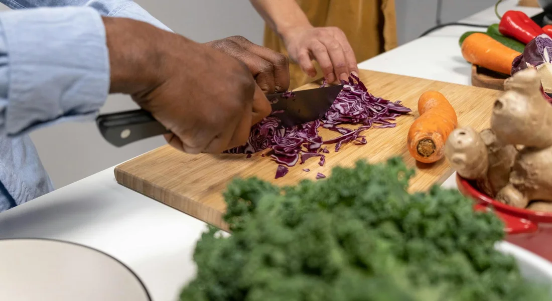 Close-up of hands chopping purple cabbage on a wooden cutting board, with carrots and kale nearby to prepare fresh veggies for a guinea pig.