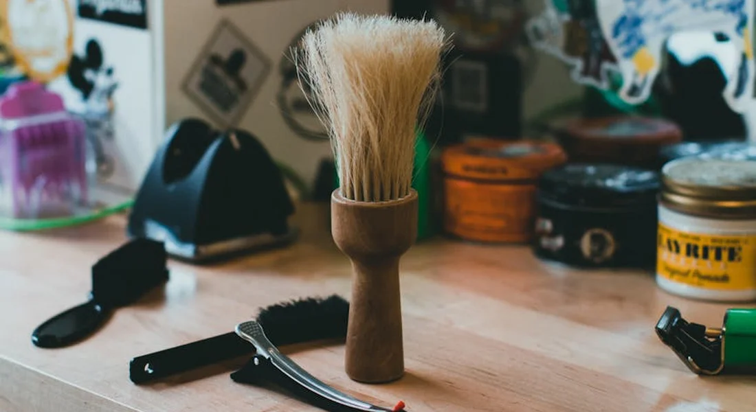 A large shaving brush stands upright on a wooden grooming table, surrounded by nail clippers and scissors, with a blurred background of jars and grooming supplies.
