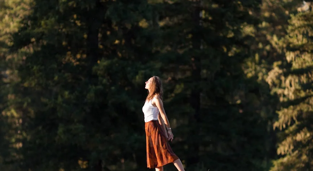 Woman walking in a sunlit forest, wearing a white top and orange skirt.