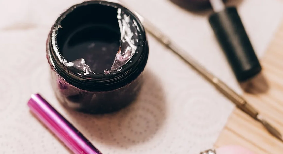 Open jar with white residues, a metal grooming tool, and a pink-tipped bottle on a paper towel, arranged as a calm manicure setup.