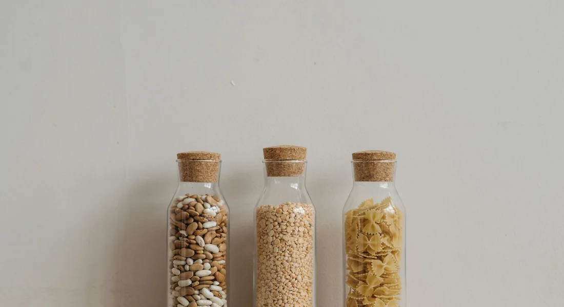 Three glass jars with cork stoppers on a light background, each jar filled with different grains or pellets.