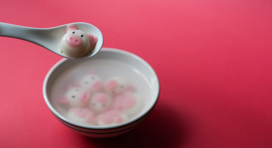 A spoon lifting a pink pig-shaped candy above a bowl containing several pink pig-shaped candies on a pink background.