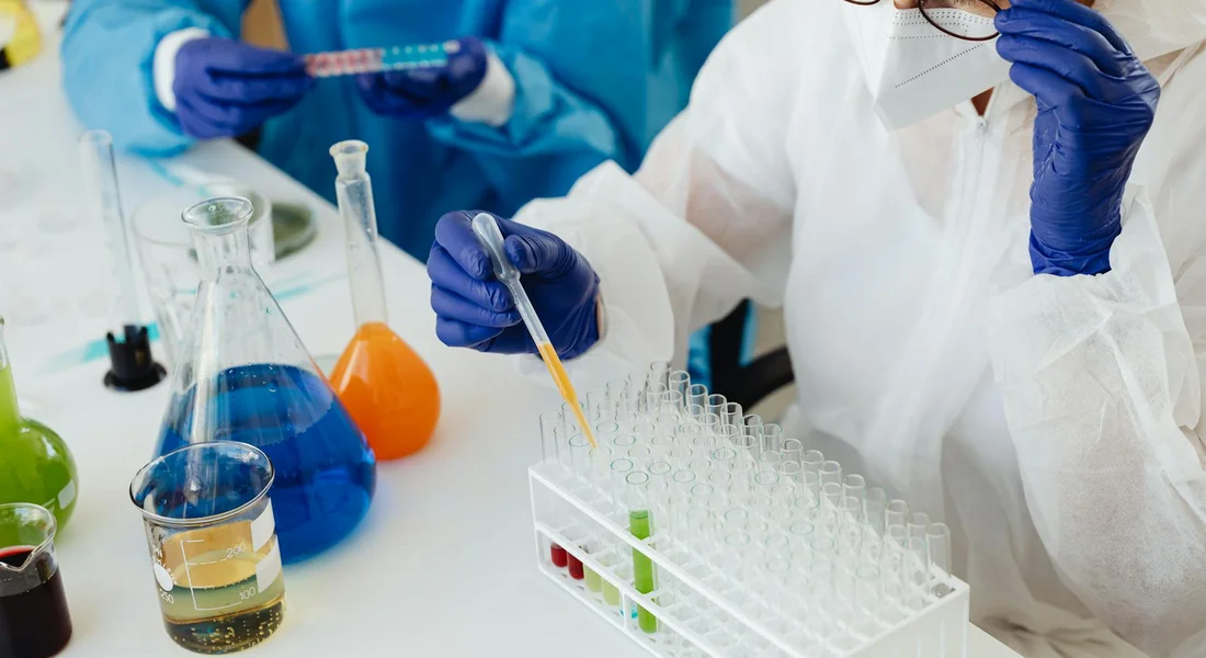 Lab scene with researchers wearing gloves and lab coats handling test tubes and a pipette over a rack of sample tubes
