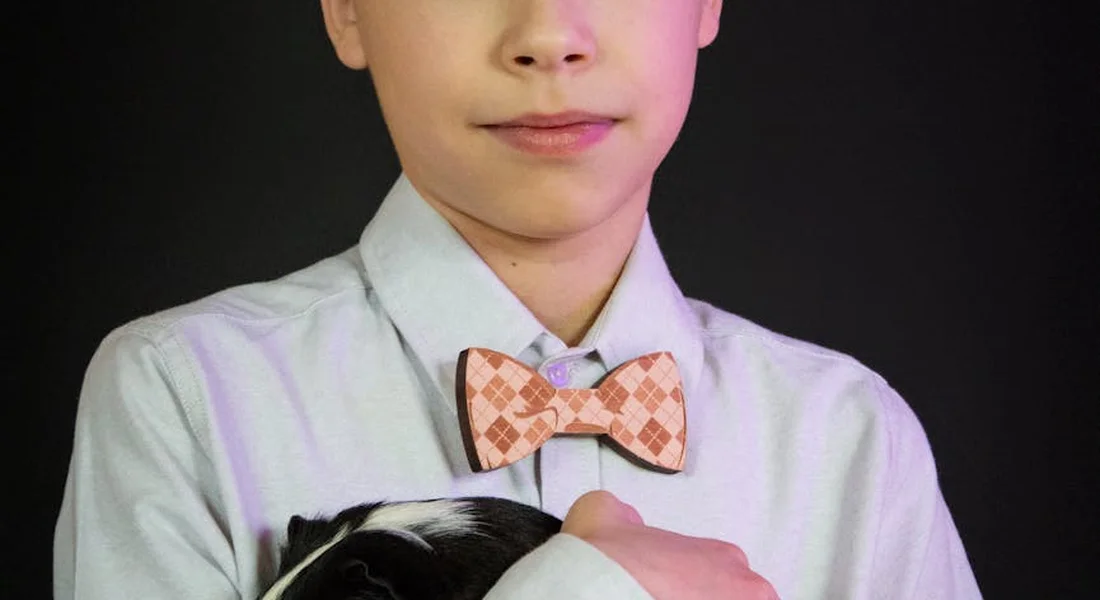 A child in a light shirt with a patterned bow tie gently holding a black and white guinea pig against a dark background.