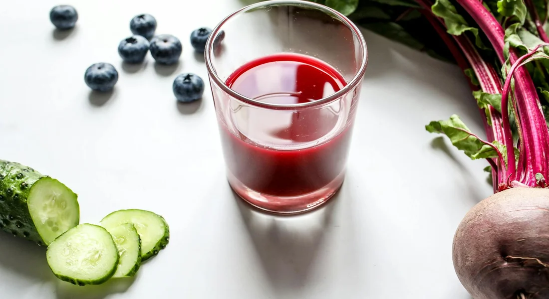 Glass of red beet juice surrounded by cucumber slices, blueberries, and beet greens on a white surface.