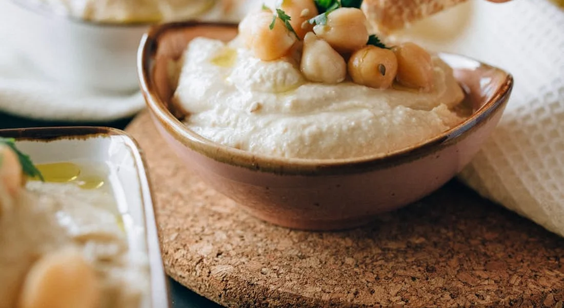 Creamy bowl of hummus with whole chickpeas on top, on a cork placemat