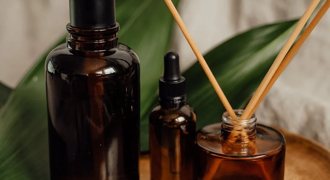 Amber glass essential oil bottles and a reed diffuser on a wooden surface with green plants in the background.