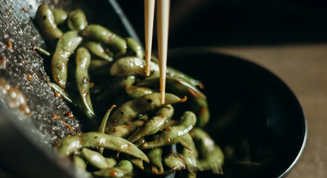 Close-up of green edamame beans in a pan being picked up with chopsticks.