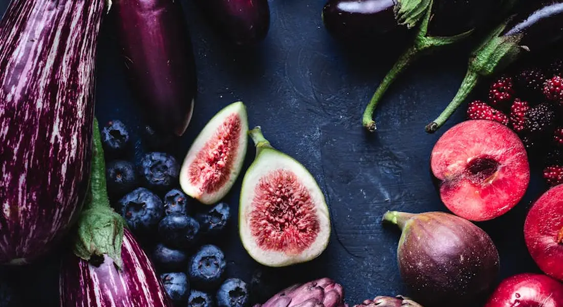 A colorful assortment of vegetables and berries on a dark surface, featuring eggplants, figs, and blueberries.