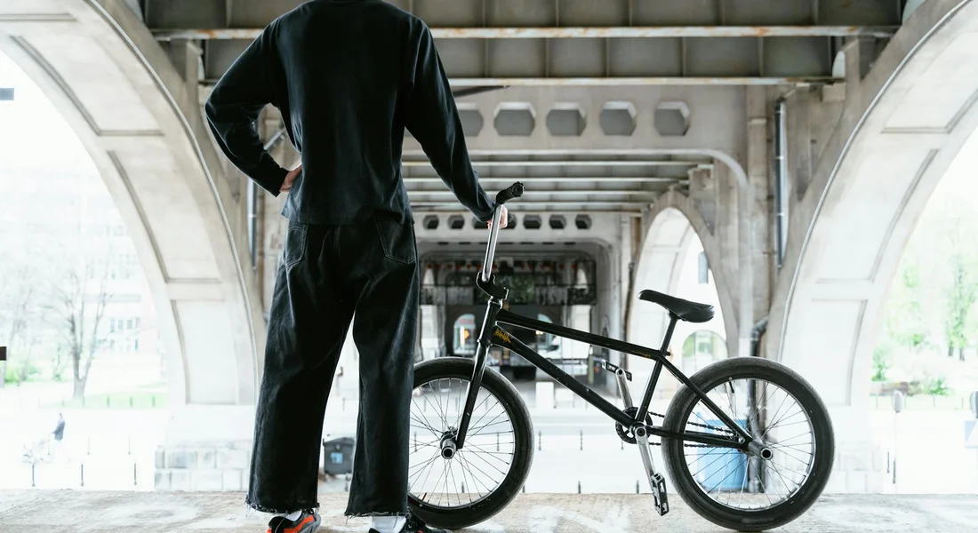 Person in dark clothing stands beside a bicycle under a covered walkway.