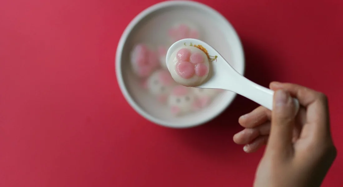 Close-up of a hand holding a white spoon with a pink, two-lobed treat above a bowl of pink and white treats on a pink background.