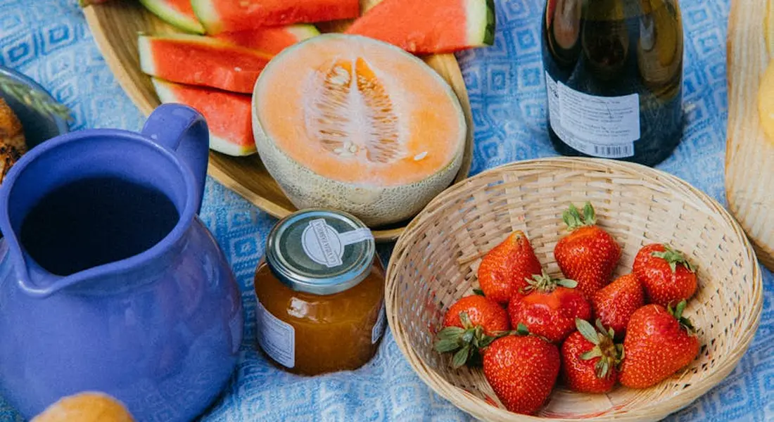 Blue patterned tablecloth with strawberries in a basket, cantaloupe slices, watermelon slices, a blue pitcher, a jar of honey, and a bottle in the background—a display highlighting foods to avoid for guinea pig safety.
