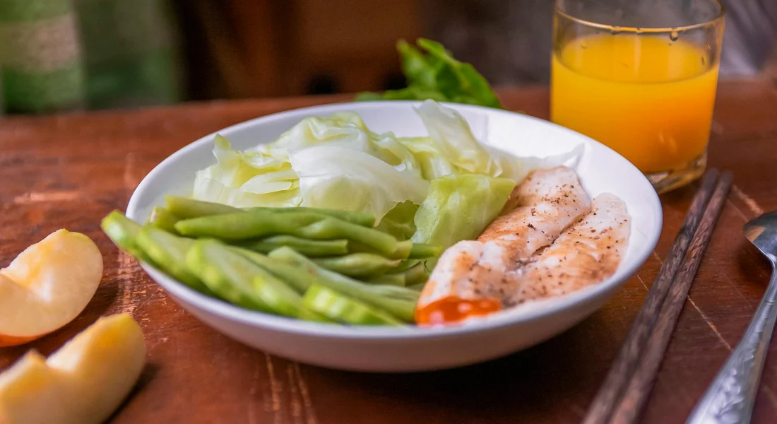 A white bowl filled with fresh green beans and lettuce on a wooden table, with a glass of orange juice and chopsticks nearby.