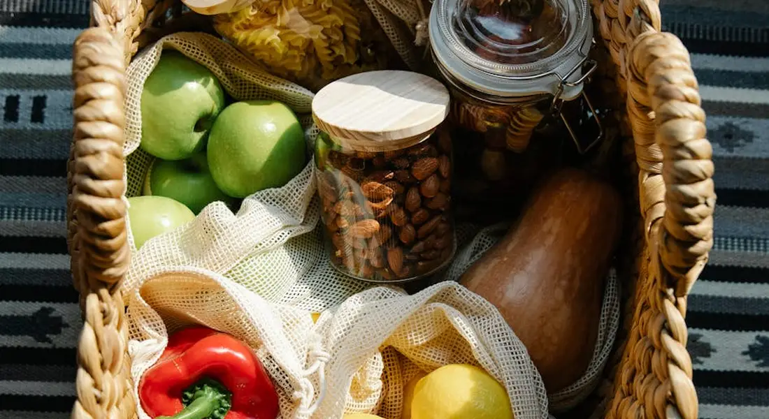 A wicker basket filled with fresh fruits—green apples, a red bell pepper, and other produce—plus jars of nuts, illustrating fruits and treats for guinea pigs.