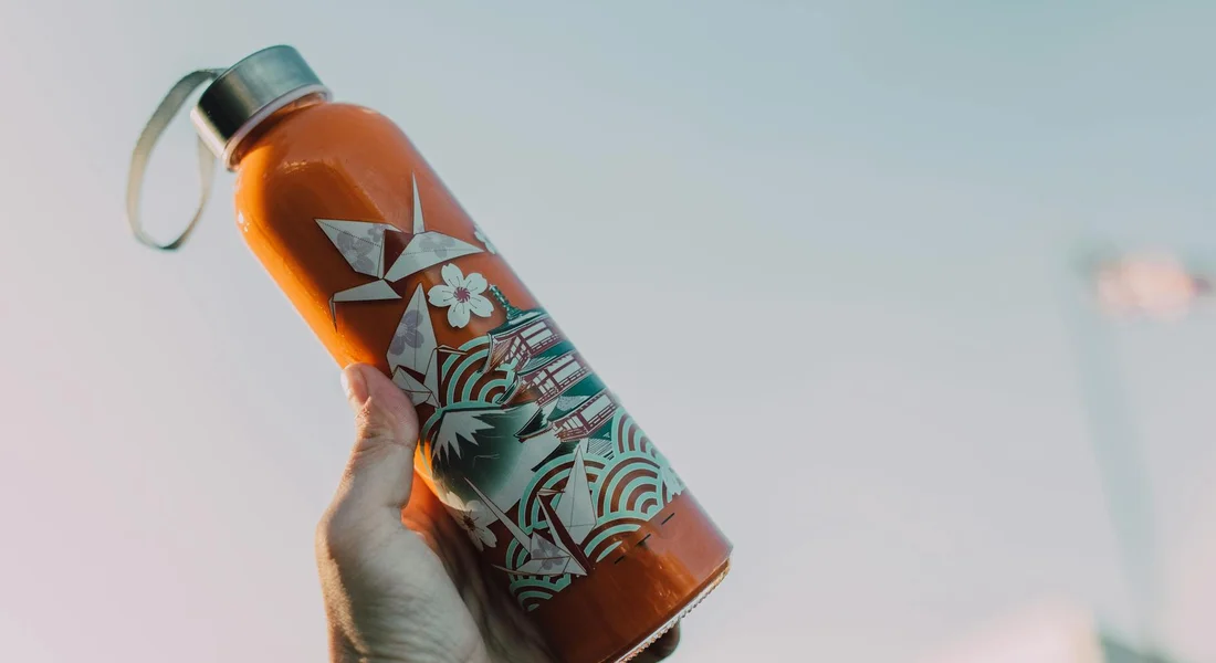 Hand holding a sports water bottle with orange liquid and decorative wrap, against a soft sky background