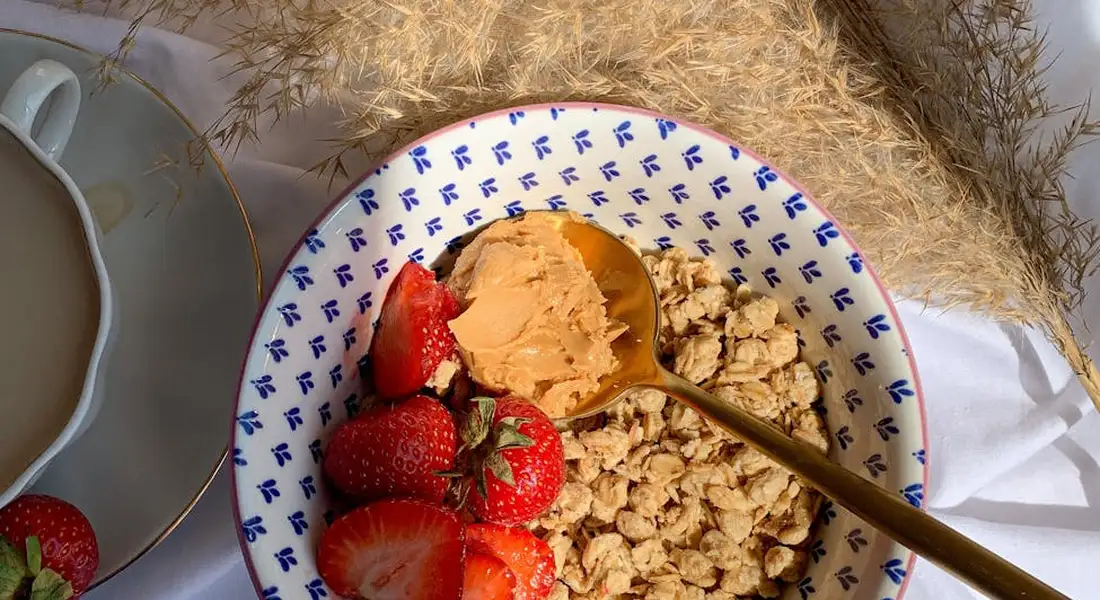 Blue-patterned bowl filled with granola and strawberry slices, with a gold spoon, beside a cup of tea on a light surface.