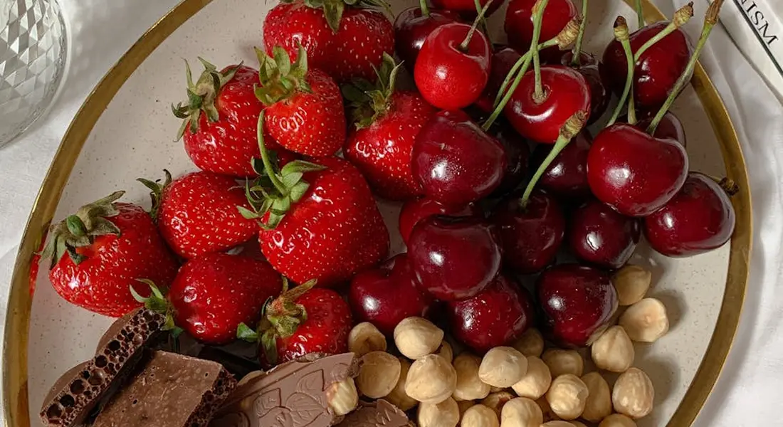 Close-up of fresh strawberries with green leaves, arranged on a plate, with a few cherries, nuts, and chocolate visible in the background.