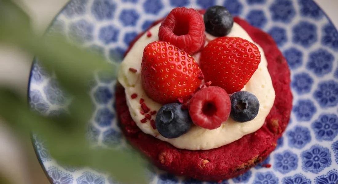 Close-up of a dessert plate with a red velvet cupcake topped with cream and fresh strawberries, raspberries, and blueberries on a blue patterned plate.