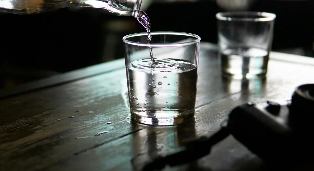 A glass of water being poured into a small glass on a wooden table, with another glass visible in the background.