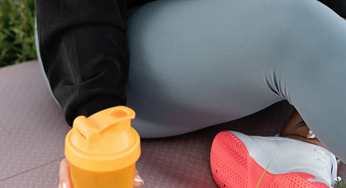 Close-up of an orange portable water bottle on a mat next to a person wearing gray leggings and pink sneakers, illustrating portable hydration options.
