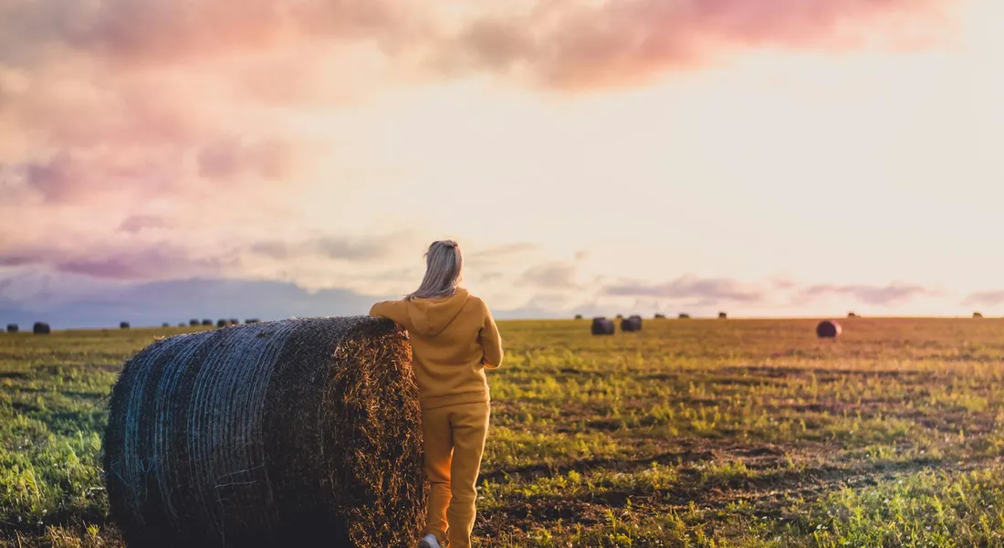 A person standing beside a large round hay bale in a sunlit field at sunset.