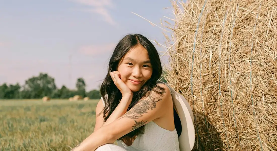 A young woman with dark hair sits beside a large hay bale in a sunlit field, smiling at the camera.