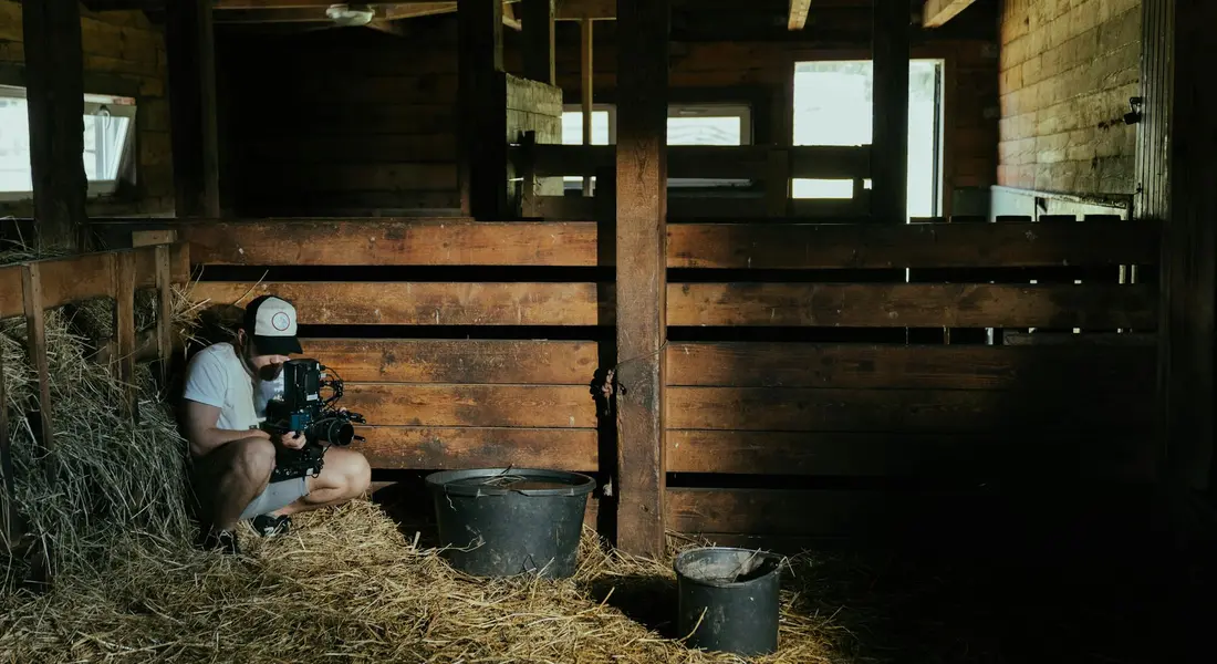 A person crouches in a dim wooden barn, filming with a camera beside stacks of hay and metal buckets.