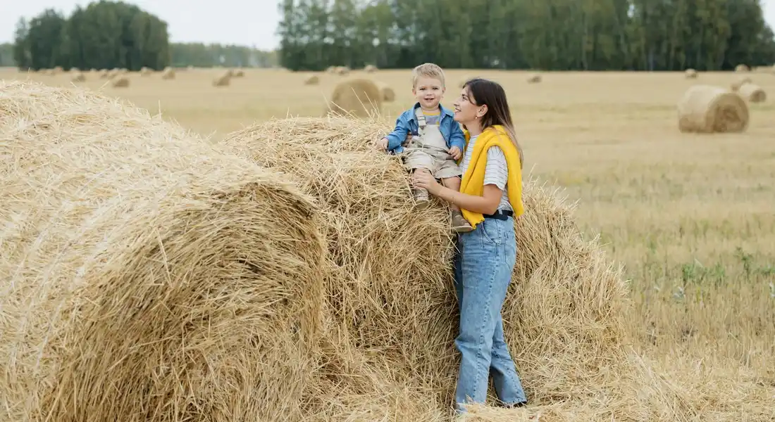 A woman and a small child stand beside a large round hay bale in a sunlit farm field, with more bales in the background.