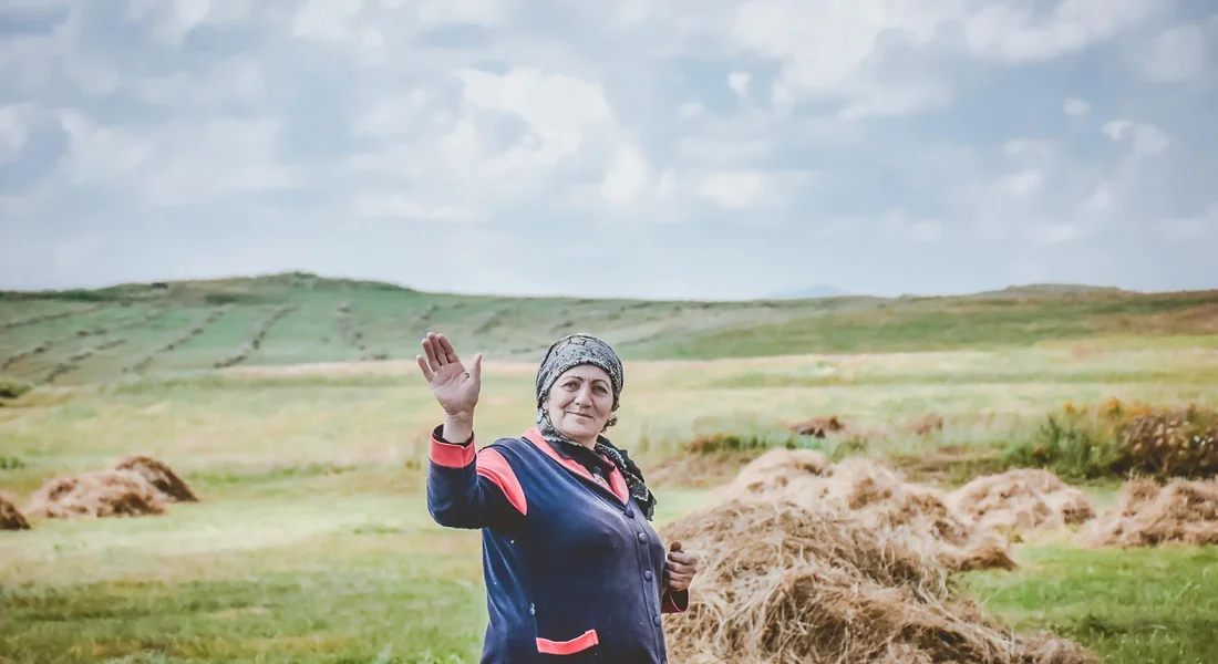 Woman standing in a rural field with hay bales in the background, waving at the camera.