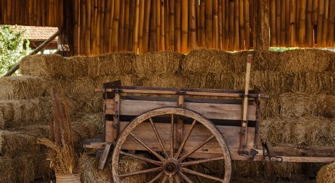Wooden cart wheel in front of stacked hay bales inside a rustic barn.