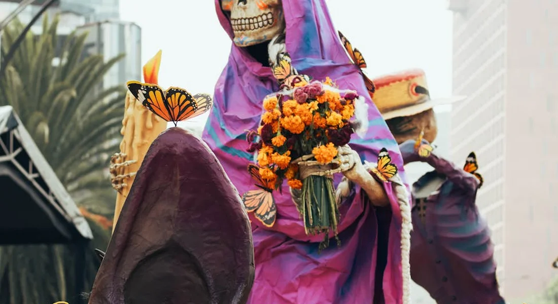 A costumed figure with a skull mask wearing a purple cloak holds a bouquet of bright orange marigold flowers.
