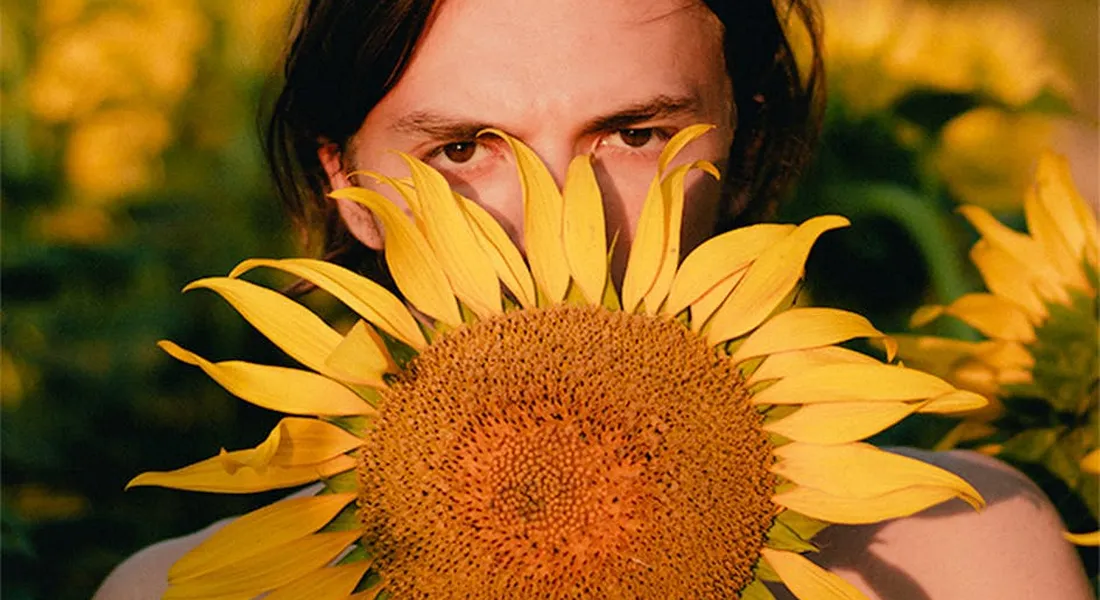 A person peeking from behind a large sunflower outdoors.