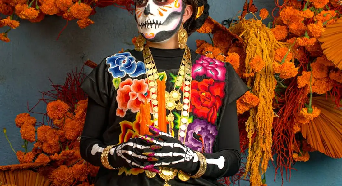 Person wearing Dia de los Muertos makeup and colorful clothing sits among bright orange marigold flowers.