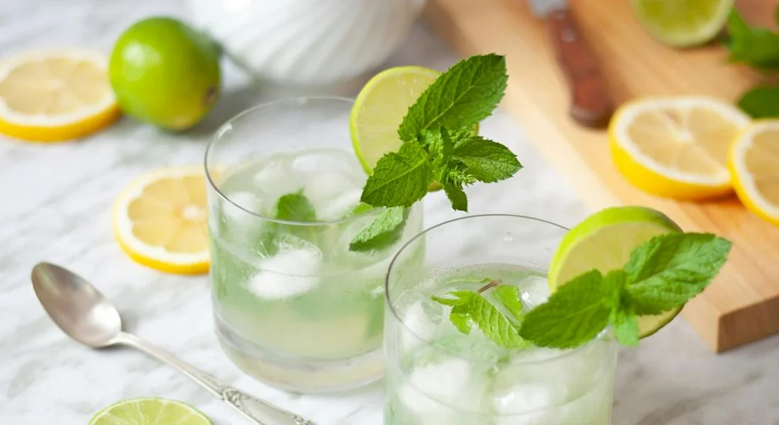 Two glasses of mint lemonade with fresh mint leaves and lime wedges on a light kitchen counter, with lemon slices in the background.