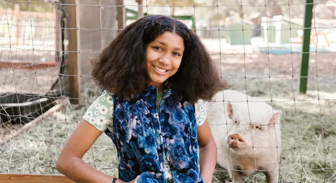 A smiling girl in a patterned shirt stands beside a white pig behind a wire fence on a farm.