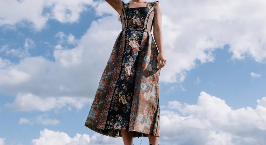 A person wearing a floral dress stands outdoors under a blue sky with clouds, photographed from a low angle.