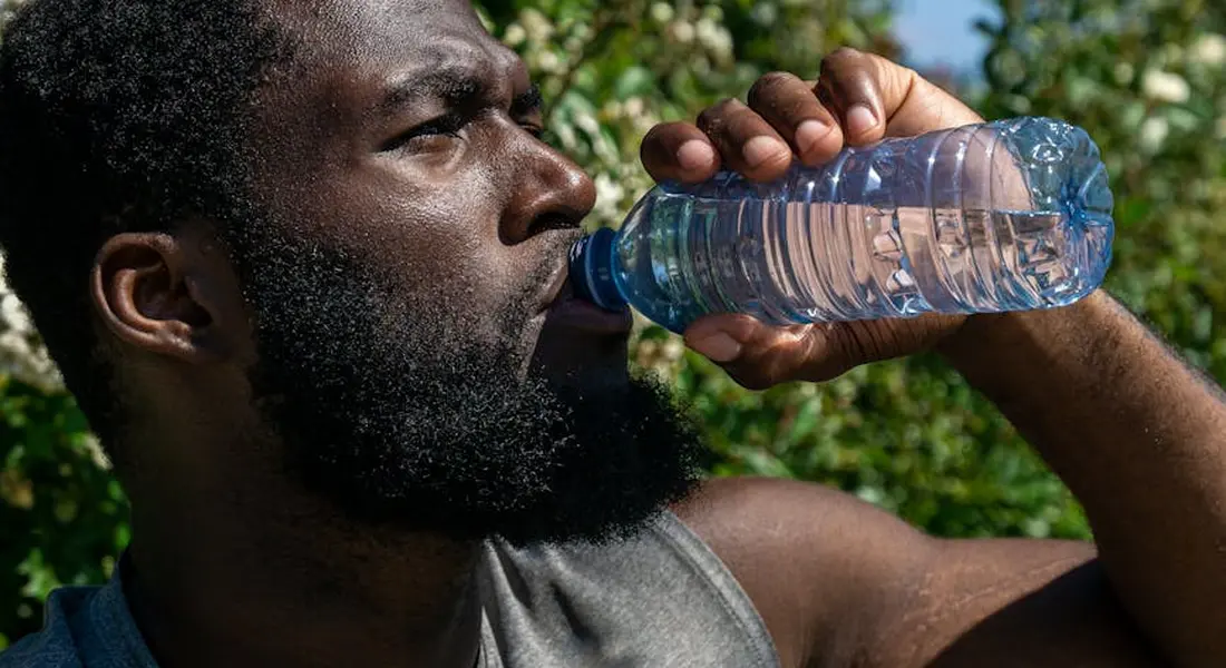 Adult man drinking from a clear plastic water bottle outdoors with green foliage in the background.