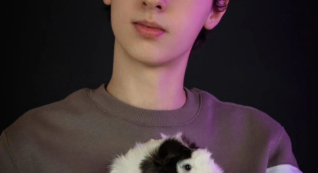 Person in a brown shirt holding a white and black guinea pig against a dark background.
